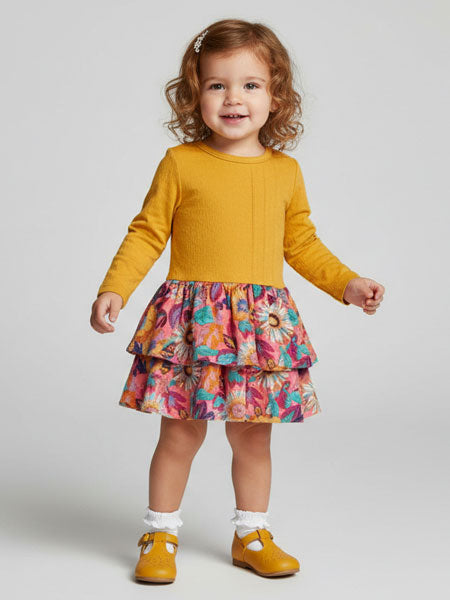 Child wearing a mustard yellow long-sleeve top and colorful floral skirt on a plain background