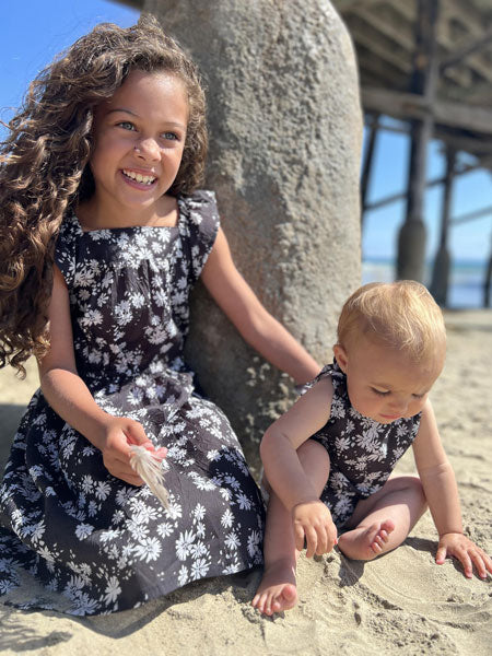 Girl modeling black daisy-printed high low dress featuring charming ruffle cap sleeves. Also pictured, baby girl in matching bubble romper.