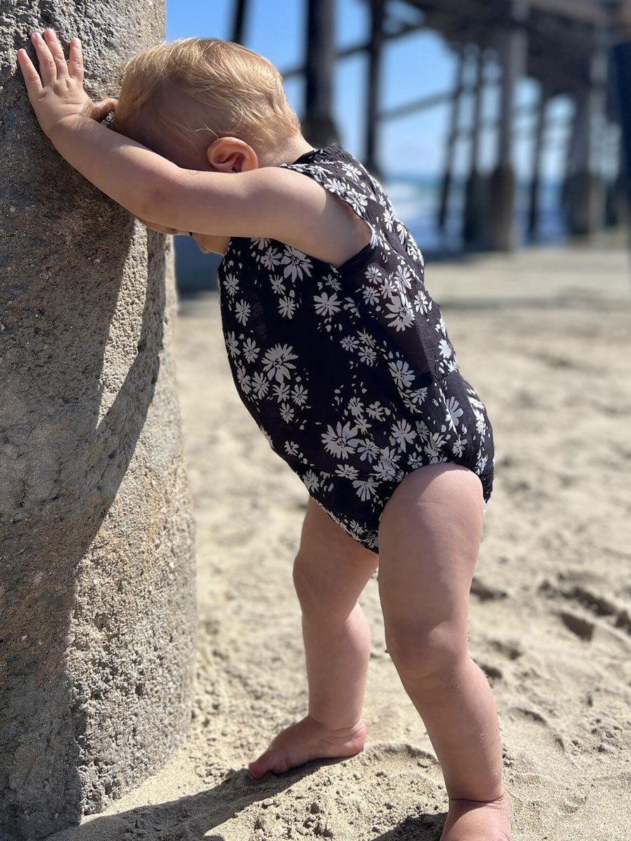 Baby girls bubble romper. Black with with daisies, sleeveless. Side view of baby girl standing in romper.