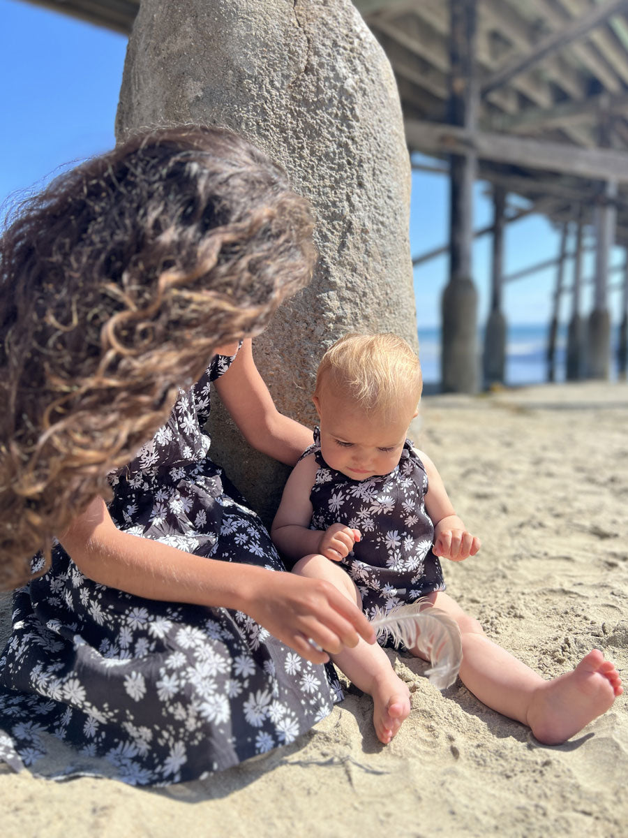 A baby girl modeling the Kate Bubble romper in black with white daisies, pictured with a girl wearing the sister matching dress, the Kim Dress by Vignette.