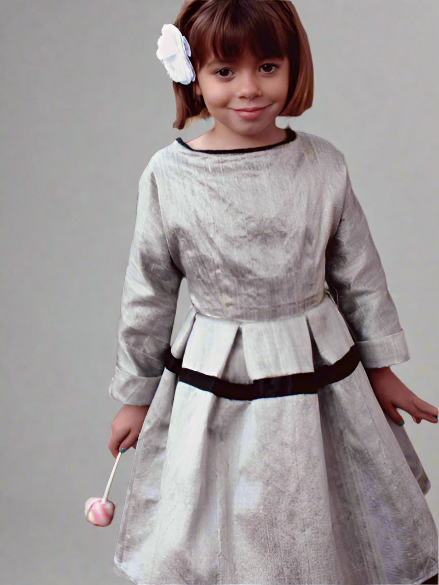 A young girl wearing a silver and black Ses Petites Mains shantung silk taffeta dress with long sleeves and etched buttons at the back neck, standing in a garden setting.