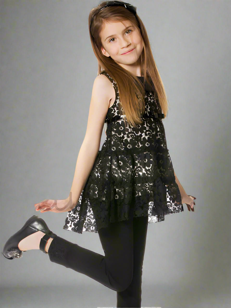 A girl wearing a black floral lace tunic top with a high-low style and a belt at the empire waist, black leggings, posing on a white background.