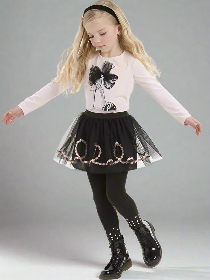 Young girl wearing a light pink ong-sleeve shirt with a black bow design, a black tulle skirt, and black shoes on a gray background.