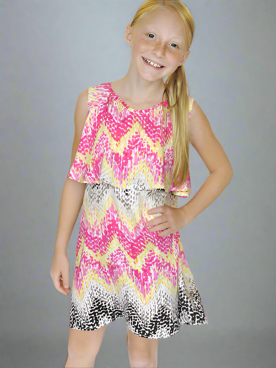 A girl wearing a multicolor A-line dress with a flowy ruffle top tier and a zig zag print. The dress has pink, taupe, chocolate, yellow, and black colors on an ivory background with a back V neckline and one button closure.