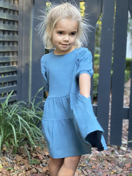 A little girl wearing teal blue cotton girls dress with her hands in side pockets. Elbow sleeves with tiered skirt.