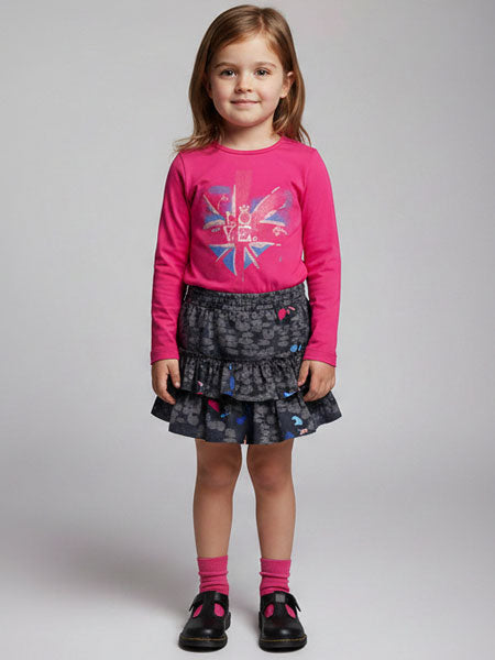 A young girl stands against a light gray studio background wearing a bright pink long-sleeve top with a colorful graphic print and a dark gray and navy patterned tiered skirt, paired with pink socks and navy blue shoes.
