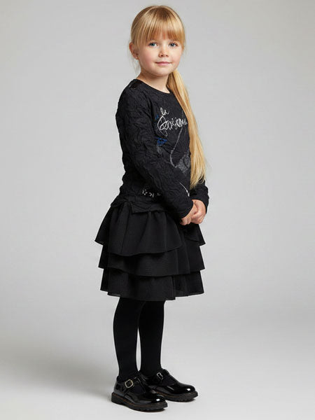 A young girl with blonde hair stands in profile wearing a black long-sleeve knit top with a graphic print, a matching black tiered ruffle skirt, black tights, and black Mary Jane shoes, against a light gray studio background.