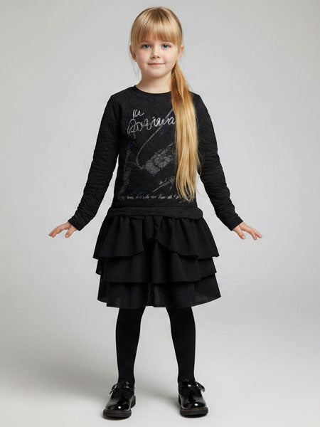 A young girl with blonde hair in a ponytail stands against a light gray studio background wearing a black long-sleeve top with a graphic print, a black tiered ruffle skirt, black tights, and black shoes.