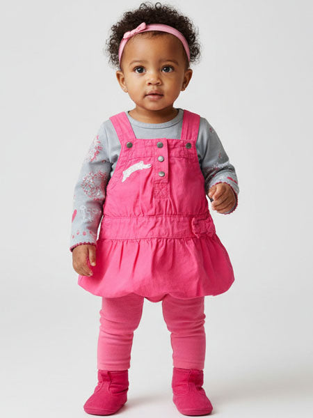 Baby wearing a pink dress overalls set with a plain background