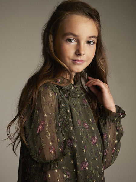 A girl shown wearing  an elegant girls dress of floral pattern on dark olive. The brand is Creamie.