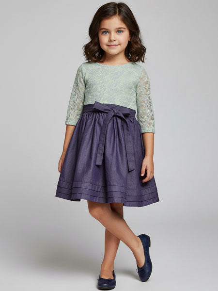 Young girl wearing a green top with lace sleeves and a purple skirt on a gray background
