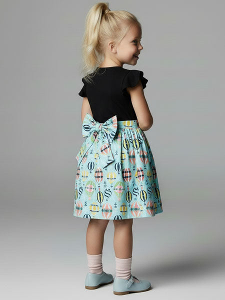 Child wearing a black top and colorful skirt with a bow on a plain background