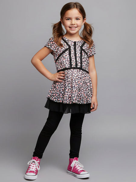 Young girl wearing a heart patterned top and black leggings with pink shoes on a gray background
