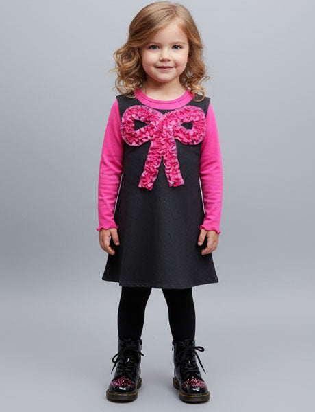 Young girl wearing a black dress with a pink bow and long-sleeve shirt on a gray background