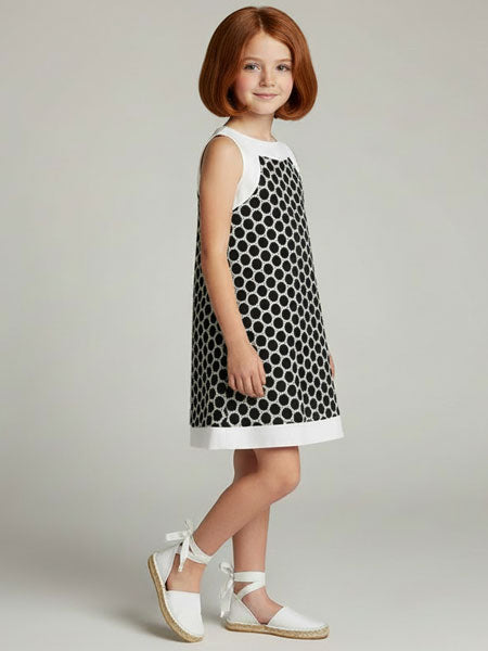 Young girl wearing a black and white polka dot dress with white shoes on a plain background