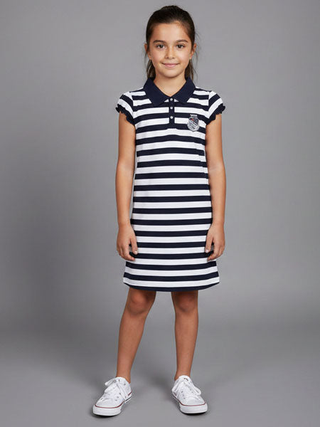 A young girl stands against a solid gray studio background wearing a navy and white horizontally striped polo dress with a small crest on the chest, paired with white sneakers.