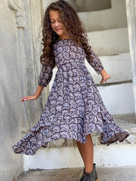 Model view, girls cotton handkerchief hem dress featuring a stunning floral block print fabric. Tiered frills on the skirt and a side tie at the waist.