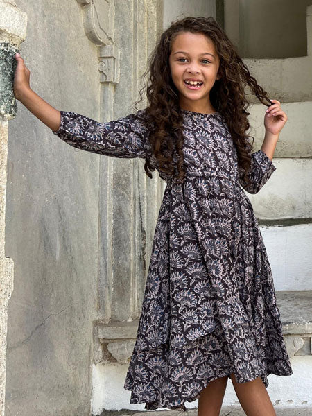 Model view, girls cotton handkerchief hem dress featuring a stunning floral block print fabric. Tiered frills on the skirt and a side tie at the waist.