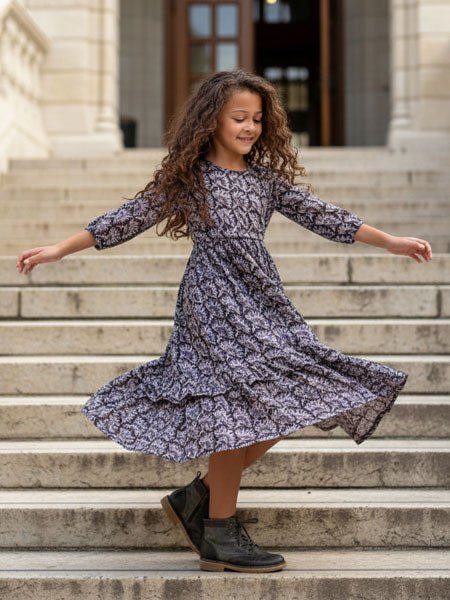 A young girl with long curly brown hair twirling on stone steps, wearing a knee-length dress with a dark gray abstract pattern and black lace-up ankle boots.
