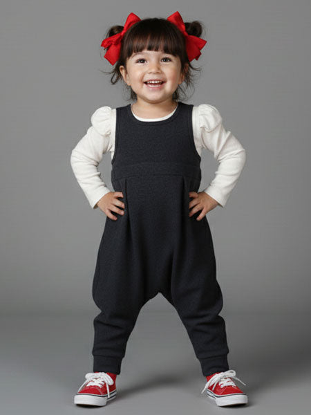 Child wearing a black dress overalls with a white shirt and red shoes on a gray background