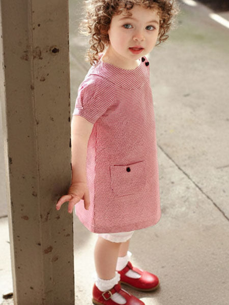 Child wearing a red houndstooth checkered dress standing on a sidewalk.