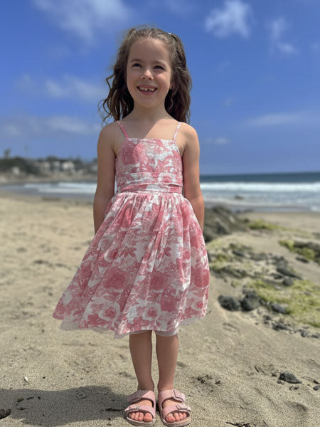 Young girl in a pink floral dress standing on a sandy beach with ocean and sky in the background.