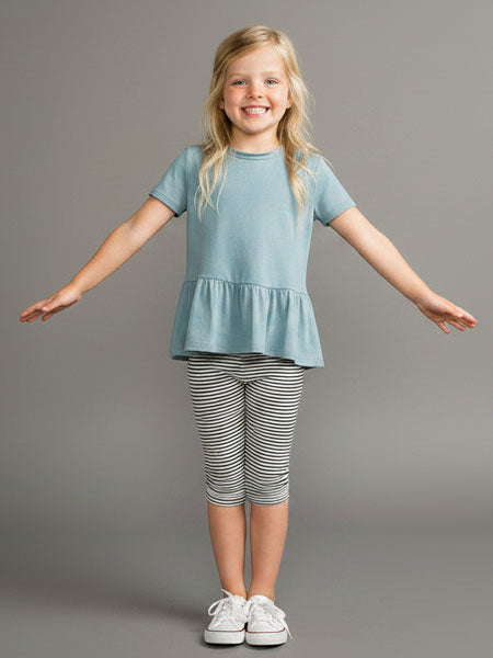 A young girl with blonde hair stands smiling with her arms outstretched, wearing a light blue short-sleeve peplum top, black and white striped capri leggings, and white sneakers against a solid gray studio background.