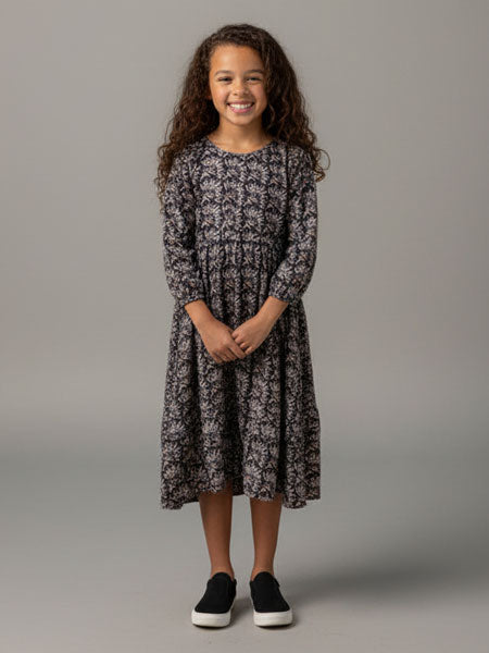 A young girl with long curly brown hair stands against a solid gray studio background wearing a long-sleeved midi dress with a dark gray and black floral pattern, paired with black slip-on shoes.