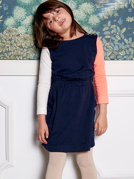 Girl modeling a color block cotton jersey dress. Body is navy with elastic gathered waist. Long sleeves, one white and one coral. Decorative gold buttons on shoulder.
