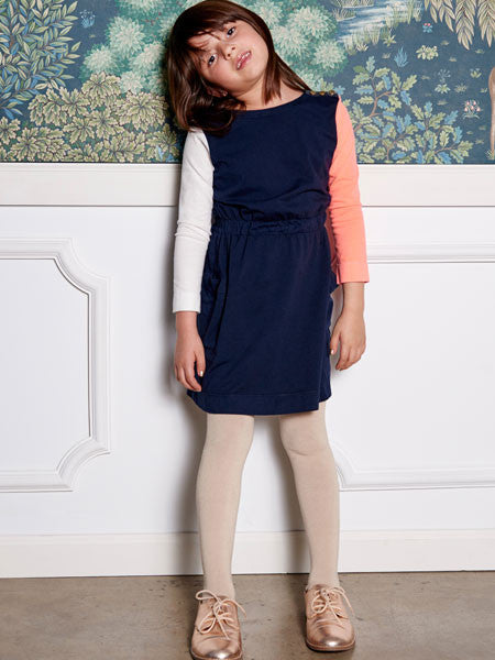 Girl modeling a color block cotton jersey dress. Body is navy with elastic gathered waist. Long sleeves, one white and one coral. Decorative gold buttons on shoulder.