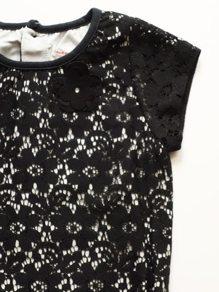 Detail view of fabric for a black lace girls party dress. Drop waist style with patent leather belt at natural waist. Short cap sleeves, white lining visible under lace.