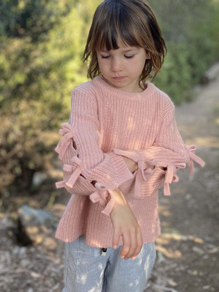 Little girl wearing pink knit crew neck sweater for girls. Three ties on sleeve outer side.